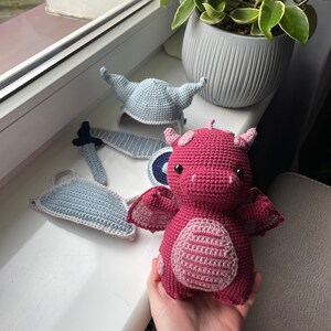 Puede incluir: Un juguete de drag&oacute;n de crochet rosa con un casco y una espada de crochet azul y blanco. El drag&oacute;n tiene un cuerpo a rayas rosa y blanco y una cola a rayas azul y blanco.