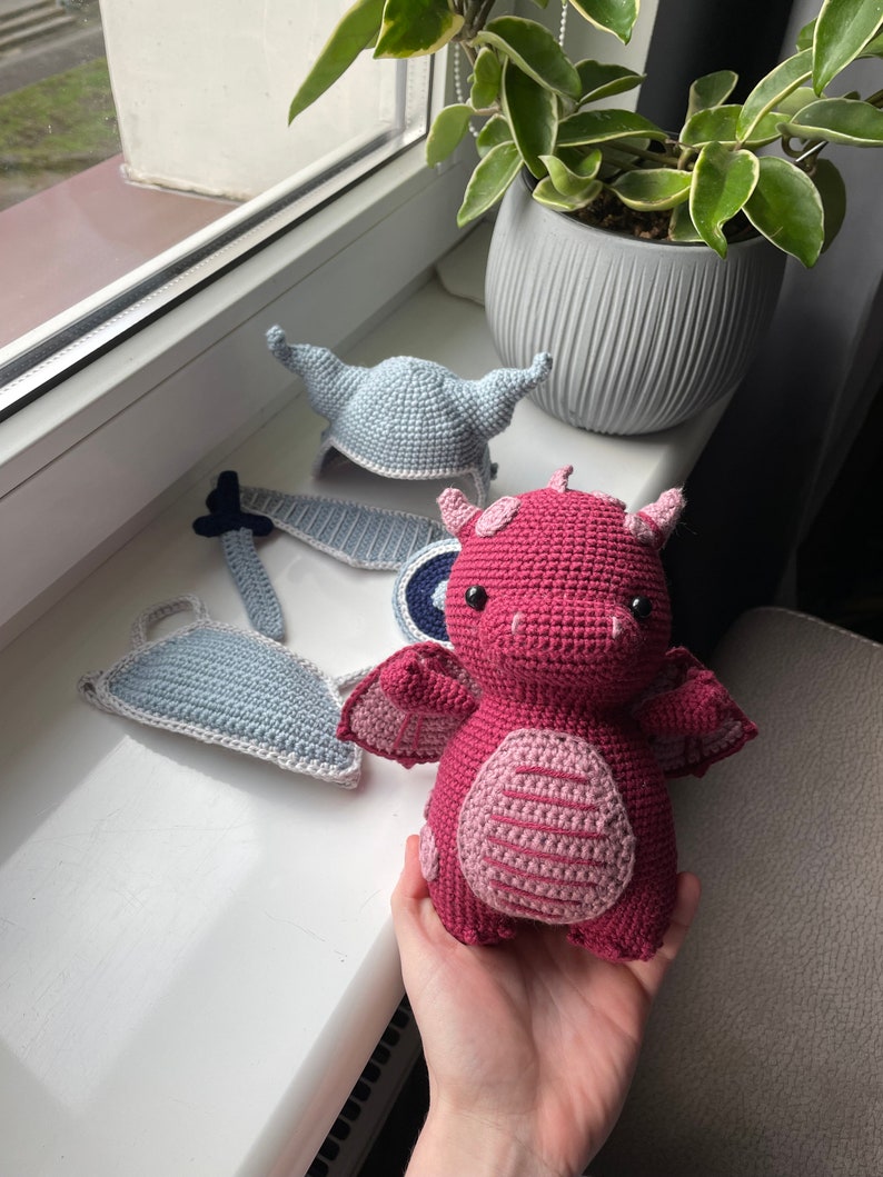 Puede incluir: Un juguete de drag&oacute;n de crochet rosa con un casco y una espada de crochet azul y blanco. El drag&oacute;n tiene un cuerpo a rayas rosa y blanco y una cola a rayas azul y blanco.