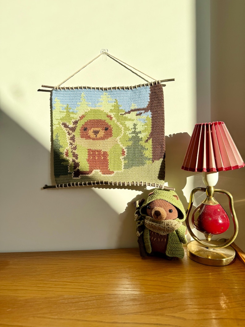 K&ouml;nnte beinhalten: Handgefertigter Wandbehang aus H&auml;kelarbeit mit einer Waldszene und einem Igel-Charakter. Das Kunstwerk ist in Gr&uuml;n-, Braun- und Blaut&ouml;nen gehalten. Ein passendes Igel-Pl&uuml;schtier und eine rote und goldene Lampe sind ebenfalls im Bild zu sehen.