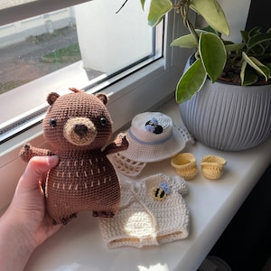 K&ouml;nnte beinhalten: Ein brauner H&auml;kelb&auml;r, der ein wei&szlig;es H&auml;keloutfit mit einem Bienendesign tr&auml;gt. Das Outfit besteht aus einem Hut, einem Shirt und einem Paar Schuhen.