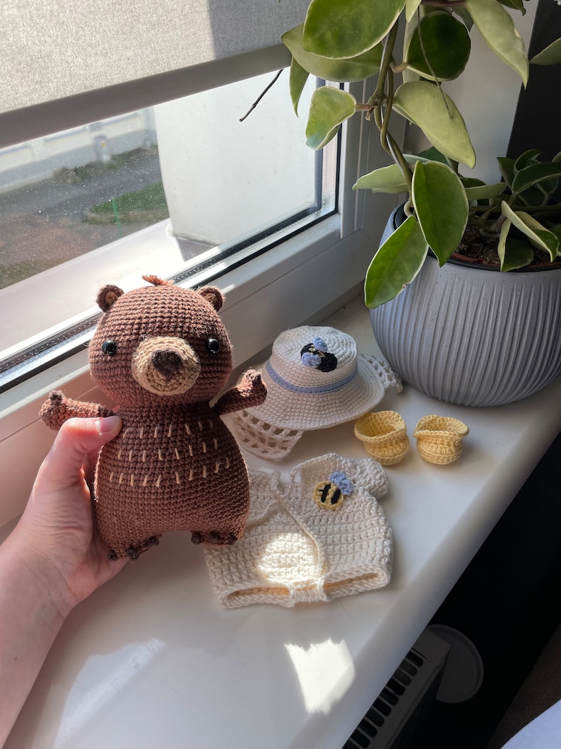 K&ouml;nnte beinhalten: Ein brauner H&auml;kelb&auml;r, der ein wei&szlig;es H&auml;keloutfit mit einem Bienendesign tr&auml;gt. Das Outfit besteht aus einem Hut, einem Shirt und einem Paar Schuhen.