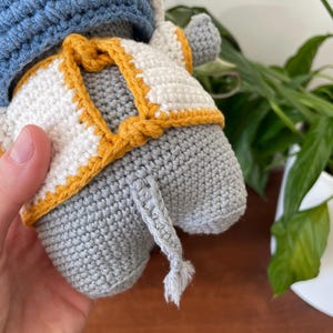 May include: A gray crocheted elephant wearing a white and yellow crocheted vest and a blue crocheted hat. The elephant has a long gray crocheted tail.