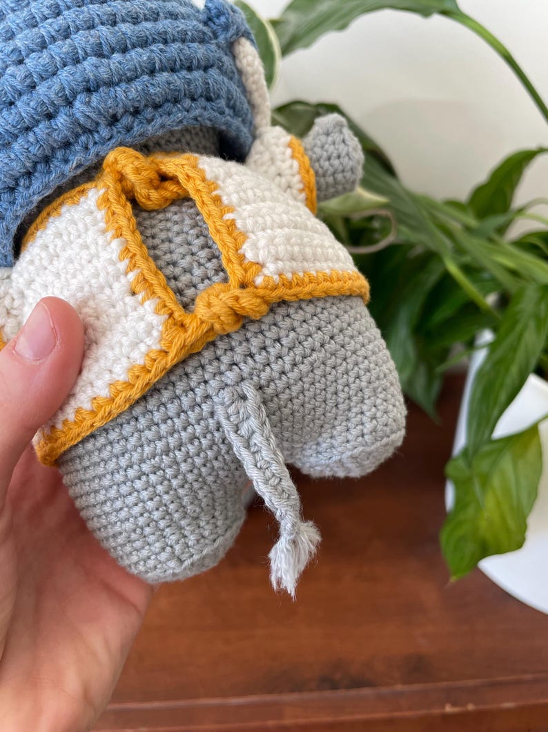 May include: A gray crocheted elephant wearing a white and yellow crocheted vest and a blue crocheted hat. The elephant has a long gray crocheted tail.