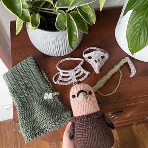 May include: A crocheted brown and pink bird with a brown sweater, a green crocheted scarf, a gray crocheted mask, and a gray crocheted collar.