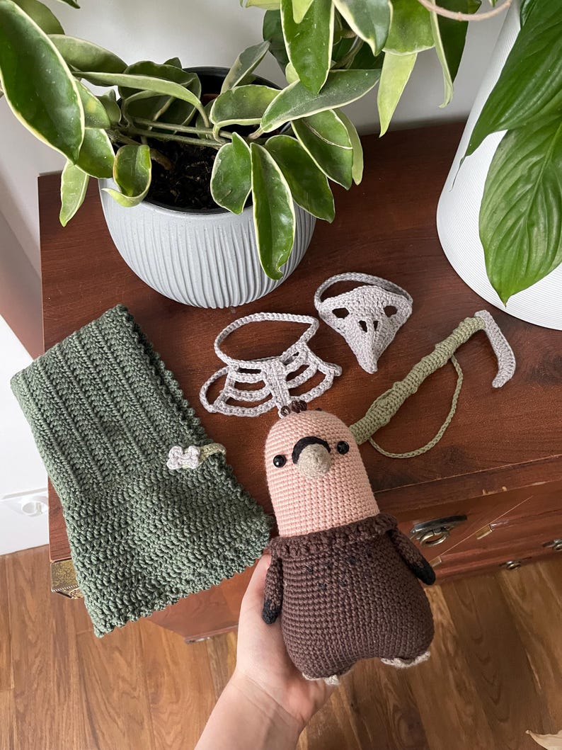May include: A crocheted brown and pink bird with a brown sweater, a green crocheted scarf, a gray crocheted mask, and a gray crocheted collar.