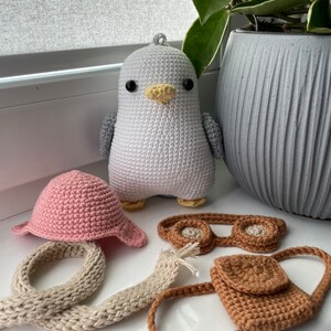 May include: A white crocheted penguin toy with black eyes and a yellow beak. The penguin is wearing a pink crocheted hat, brown crocheted goggles, a brown crocheted bag, and a beige crocheted scarf.
