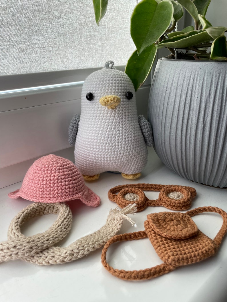 May include: A white crocheted penguin toy with black eyes and a yellow beak. The penguin is wearing a pink crocheted hat, brown crocheted goggles, a brown crocheted bag, and a beige crocheted scarf.