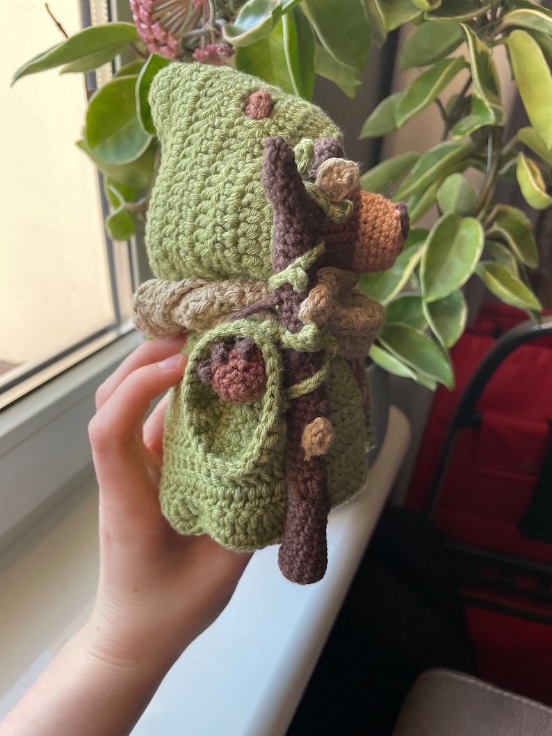 May include: A crocheted green gnome doll with a brown stick and brown leaves. The gnome is wearing a green hat and has a brown beard.