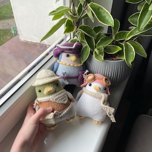 Può includere: Tre giocattoli di pinguino all'uncinetto, ognuno con un abbigliamento diverso. Un pinguino indossa un cappello viola e blu, un altro indossa un cappello verde e un gilet marrone, e il terzo indossa un cappello rosa con occhiali e una sciarpa bianca.