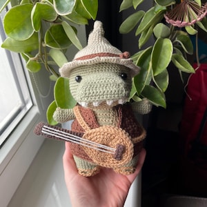 Puede incluir: Un dinosaurio de crochet verde que lleva un sombrero marrón y sostiene un laúd marrón.