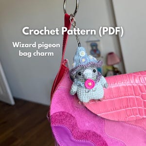 May include: A crochet pattern PDF for a wizard pigeon bag charm. The charm is a light blue crocheted pigeon wearing a pointed hat with white buttons and a purple brim, featuring a bright pink button on its chest, attached to a pink handbag.