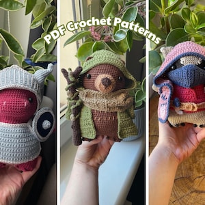 May include: Three crocheted fantasy creatures are shown. The first is a red creature in a silver helmet and armour, holding a sword and shield. The second is a brown bear-like creature in a green hooded cloak. The third is a tan creature in a pink and red hooded outfit.