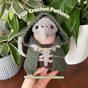 May include: Crochet pattern for a Necromancer Vulture amigurumi. The amigurumi is wearing a green hooded robe and has a pink beak and head. The pattern is available as a PDF download.
