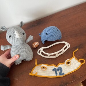 May include: A gray crocheted rhinoceros plush toy, a blue crocheted helmet, a white crocheted football helmet face mask, a white crocheted jersey with a yellow trim and the number 12 in blue, and a small orange crocheted football.