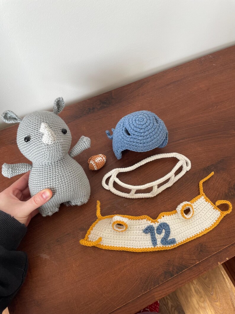 May include: A gray crocheted rhinoceros plush toy, a blue crocheted helmet, a white crocheted football helmet face mask, a white crocheted jersey with a yellow trim and the number 12 in blue, and a small orange crocheted football.