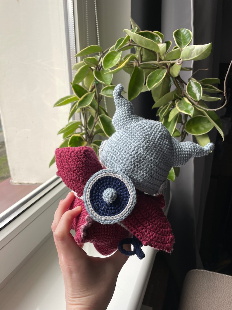 Puede incluir: Un juguete de peluche tejido a crochet con un casco gris, alas rojas y un escudo azul y blanco. El juguete se sostiene en una mano contra una ventana.