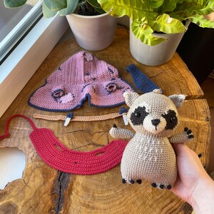 May include: A handmade crochet raccoon doll with black and white accents, a red crochet piece, and a pink and blue crochet moth. The items are displayed on a wooden surface near potted plants.