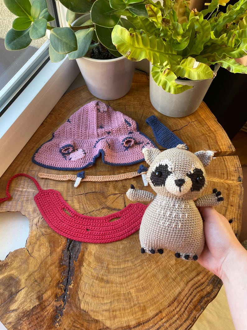 May include: A handmade crochet raccoon doll with black and white accents, a red crochet piece, and a pink and blue crochet moth. The items are displayed on a wooden surface near potted plants.