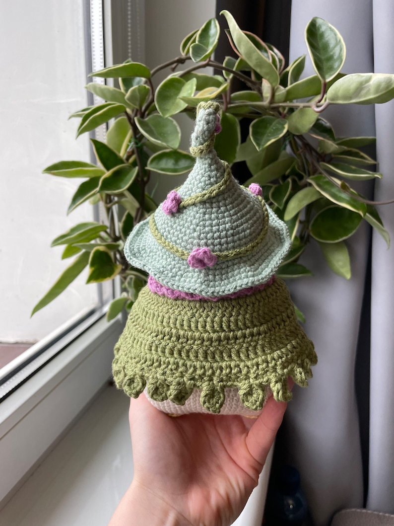 May include: A crocheted gnome doll with a green hat, pink flowers, and a green skirt.