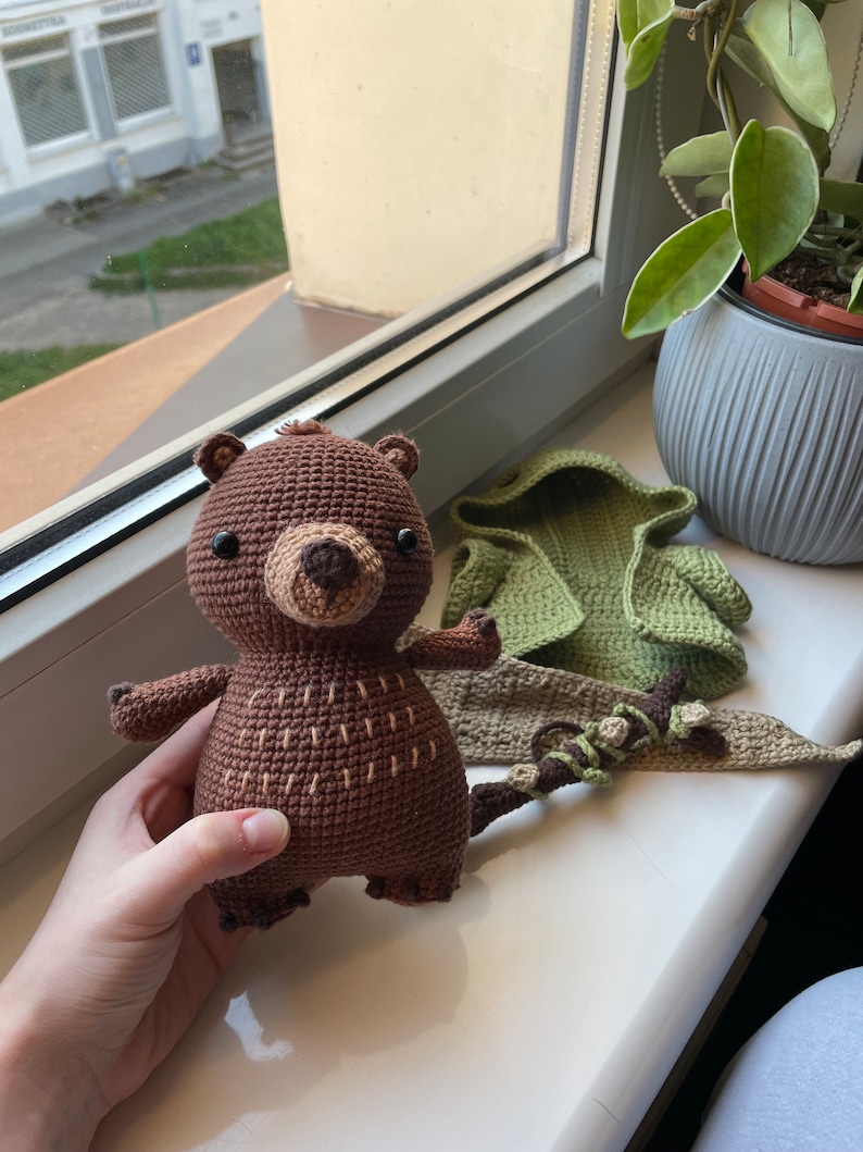 Puede incluir: Un oso de crochet marr&oacute;n con ojos negros y una nariz marr&oacute;n. El oso lleva una chaqueta de crochet verde y tiene una cola de crochet verde.
