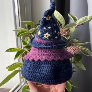 May include: A handmade crochet witch hat in shades of navy blue and purple. The hat features embroidered yellow stars and a decorative purple brim. The base of the hat is a rounded shape, and the top is a pointed spiral.