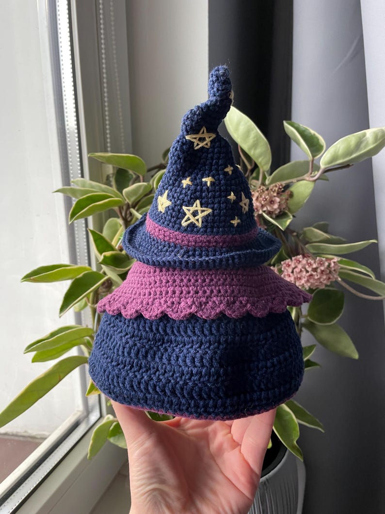 May include: A handmade crochet witch hat in shades of navy blue and purple. The hat features embroidered yellow stars and a decorative purple brim. The base of the hat is a rounded shape, and the top is a pointed spiral.