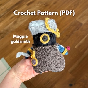 Może przedstawiać: Szydełkowa figurka amigurumi z czarną głową, żółtą obwódką oka, szarym kapeluszem i brązowym fartuchem. Figurka ma niebieskie rękawy, żółte akcenty i trzyma małe narzędzie. Tekst: Crochet Pattern (PDF) Magpie goldsmith.