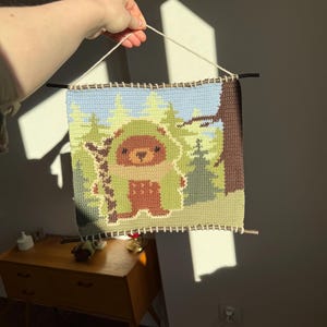K&ouml;nnte beinhalten: Handgefertigter Wandbehang aus H&auml;kelarbeit mit einem B&auml;ren in einer Waldszene. Das Kunstwerk verwendet Braun-, Gr&uuml;n- und Blaut&ouml;ne. Das St&uuml;ck ist rechteckig, mit einer hellen Schnur zum Aufh&auml;ngen und dunklen St&auml;ben oben und unten.