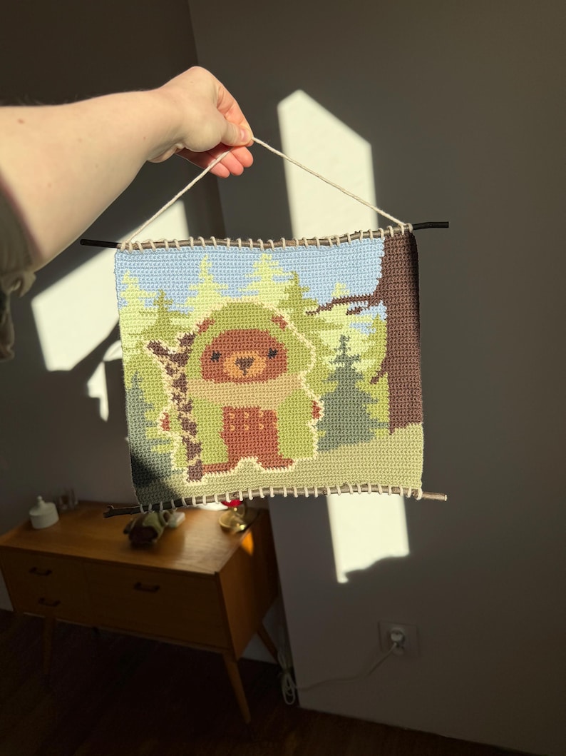 K&ouml;nnte beinhalten: Handgefertigter Wandbehang aus H&auml;kelarbeit mit einem B&auml;ren in einer Waldszene. Das Kunstwerk verwendet Braun-, Gr&uuml;n- und Blaut&ouml;ne. Das St&uuml;ck ist rechteckig, mit einer hellen Schnur zum Aufh&auml;ngen und dunklen St&auml;ben oben und unten.