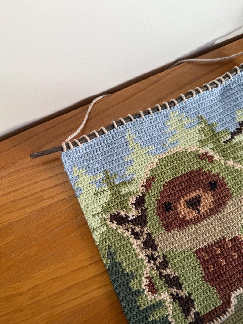 K&ouml;nnte beinhalten: Ein unfertiges H&auml;kelprojekt mit einer Waldszene und einem B&auml;ren. Das St&uuml;ck ist haupts&auml;chlich gr&uuml;n, blau und braun, mit einem hellblauen Rand. Ein Holzstab und Garn sind oben befestigt.