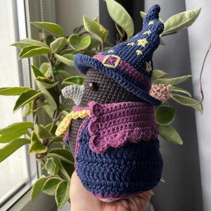 May include: A crocheted stuffed bird wearing a purple and blue wizard hat with yellow stars. The bird is wearing a purple and blue cape with a yellow trim.