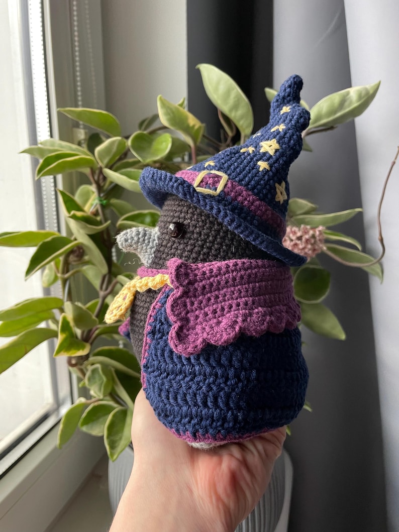 May include: A crocheted stuffed bird wearing a purple and blue wizard hat with yellow stars. The bird is wearing a purple and blue cape with a yellow trim.