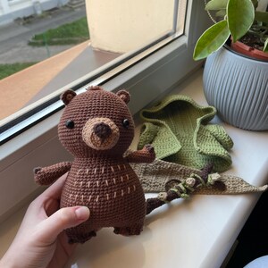 May include: A handmade brown crochet bear with a tan snout and small black eyes. The bear has small arms and legs, and a tail. Beside the bear are a green crochet hood and a brown and green crochet accessory.