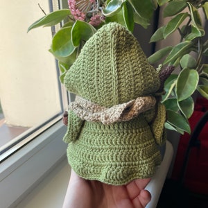 Puede incluir: Mu&ntilde;eca amigurumi de crochet verde con una capucha de crochet marr&oacute;n y un cuello de crochet marr&oacute;n. La mu&ntilde;eca lleva un vestido de crochet verde.