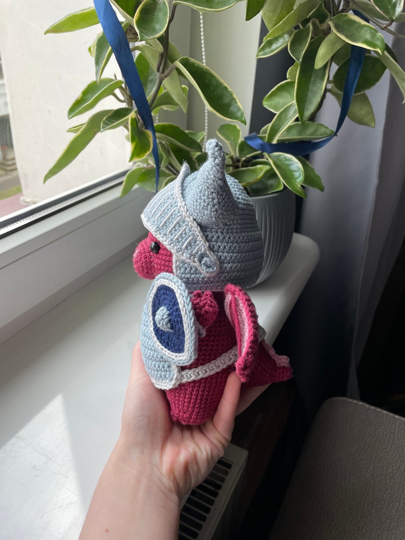 Puede incluir: Un drag&oacute;n de crochet con un casco y un escudo azul y blanco. El drag&oacute;n es rosa y blanco y tiene una cola peque&ntilde;a y puntiaguda.