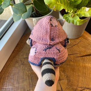 May include: A handmade crochet raccoon doll in pink, gray, and black. The doll has a hooded jacket with blue stitching and a striped tail. The doll is made of yarn and is a soft toy.