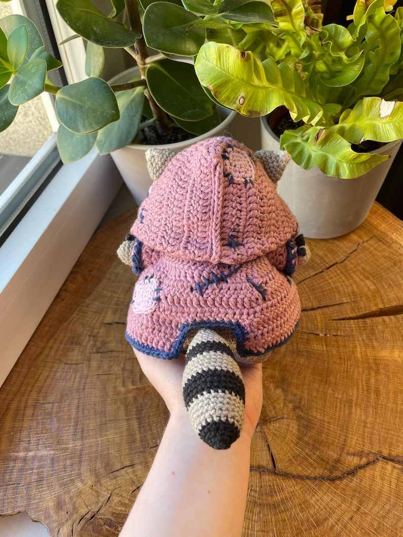 May include: A handmade crochet raccoon doll in pink, gray, and black. The doll has a hooded jacket with blue stitching and a striped tail. The doll is made of yarn and is a soft toy.