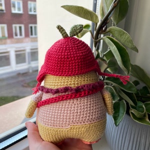 May include: A crocheted amigurumi toy shaped like a person wearing a red crocheted hat with a green leaf on top. The toy is wearing a pink and yellow crocheted outfit with a red and pink crocheted scarf.