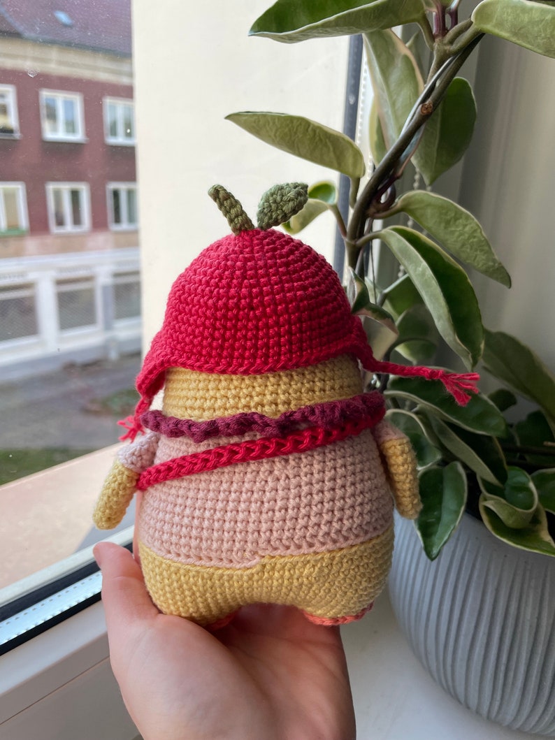 May include: A crocheted amigurumi toy shaped like a person wearing a red crocheted hat with a green leaf on top. The toy is wearing a pink and yellow crocheted outfit with a red and pink crocheted scarf.
