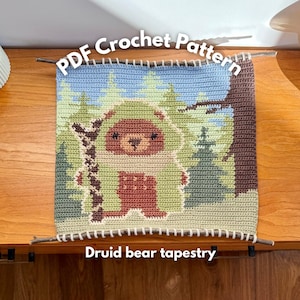 Puede incluir: Un tapiz de ganchillo que representa un oso en un bosque. El tapiz utiliza tonos verdes, marrones y azules, con el texto "PDF Crochet Pattern" y "Druid bear tapestry". El tapiz se exhibe sobre una superficie de madera.