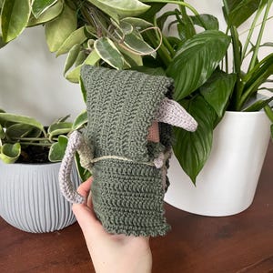 May include: A crocheted green hooded figure with a pink face and tail. The figure is being held by a hand.