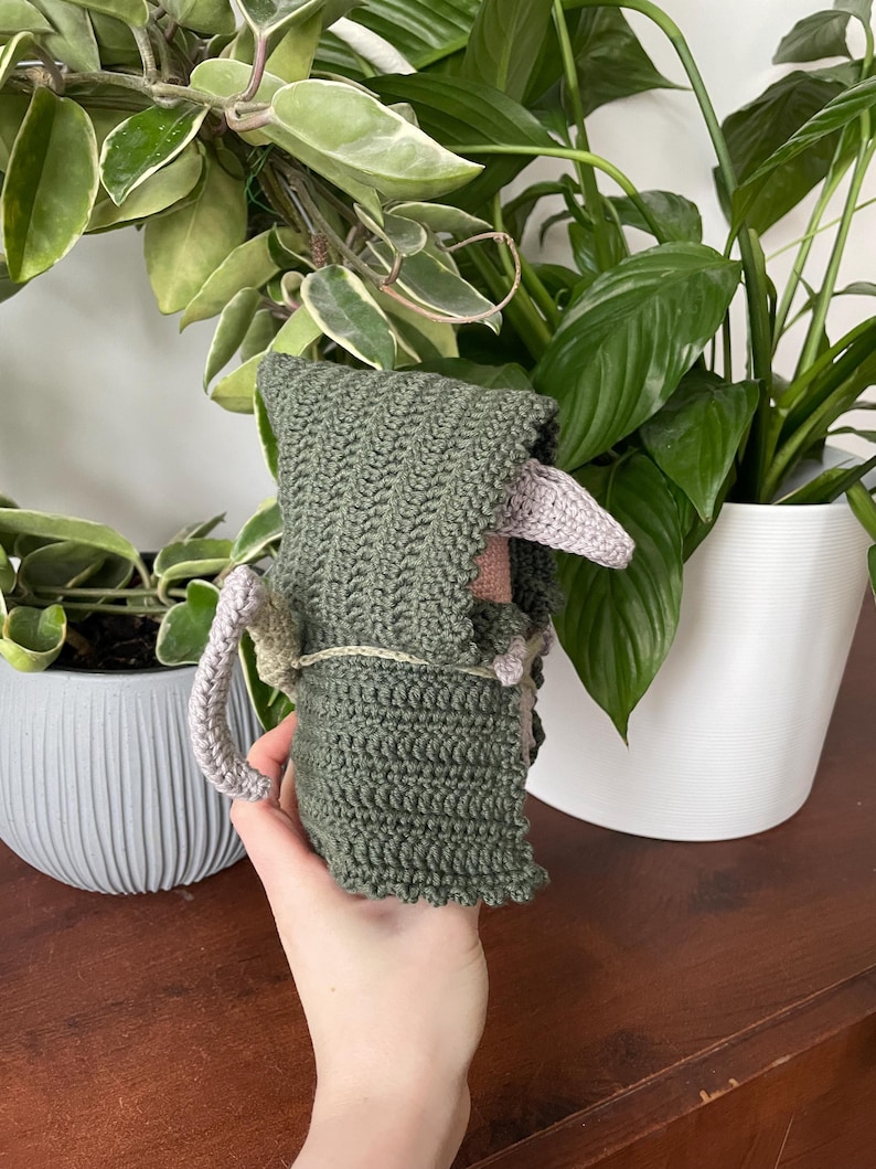 May include: A crocheted green hooded figure with a pink face and tail. The figure is being held by a hand.