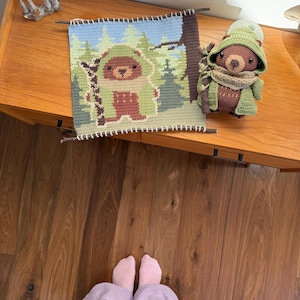 K&ouml;nnte beinhalten: Ein handgefertigter Wandbehang aus H&auml;kelarbeit und ein Stofftier in Form eines B&auml;ren. Der Wandbehang zeigt einen B&auml;ren in einer Waldszene mit blauen, gr&uuml;nen und braunen Farben. Das B&auml;r-Spielzeug tr&auml;gt einen gr&uuml;nen Hut, Schal und Mantel. Die Artikel befinden sich auf einer Holzoberfl&auml;che.