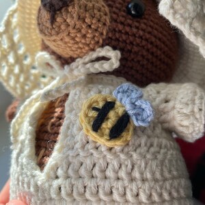 K&ouml;nnte beinhalten: Ein geh&auml;kelter brauner B&auml;r, der ein wei&szlig;es H&auml;kelkleid mit einer Bienenapplikation tr&auml;gt. Die Biene ist gelb mit schwarzen Streifen und hat eine blaue Blume oben drauf.