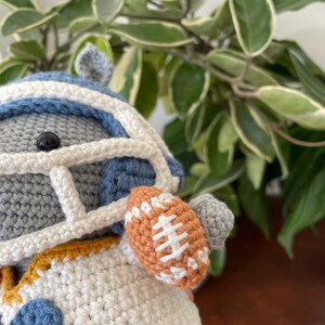 May include: A gray crocheted stuffed animal wearing a blue and white football helmet, a white jersey with a blue number "2" and a yellow trim, and holding a brown football.