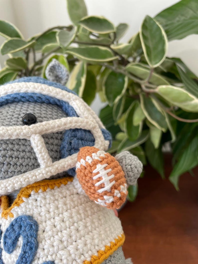 May include: A gray crocheted stuffed animal wearing a blue and white football helmet, a white jersey with a blue number "2" and a yellow trim, and holding a brown football.