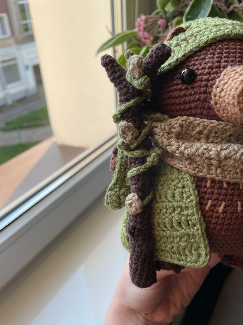 May include: A crocheted stuffed toy with brown fur, a green hat, and a brown stick in its hand. The toy is wearing a brown and beige scarf.