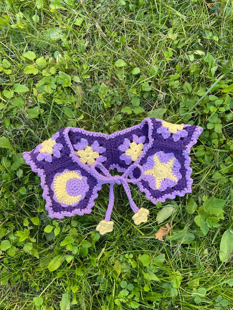 Crochet granny pentagon collar made in light purple, dark purple and yellow color scheme laying on green grass. Collar consist of 6 granny pentagons, 4 regular, 1 star and 1 moon. It is tied using light purple ties with little stars at the end.