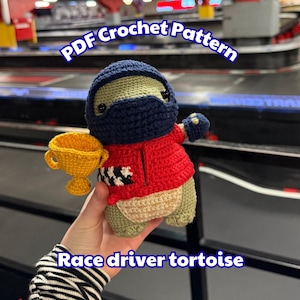 May include: A crocheted race driver tortoise doll wearing a navy blue helmet and mask, a red jacket, and holding a gold trophy. The doll has a beige body and green legs. The text "PDF Crochet Pattern" and "Race driver tortoise" are visible.