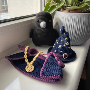 May include: A crocheted black bird with a white beak, a blue and purple crocheted robe with a gold chain, and a blue crocheted hat with yellow stars.